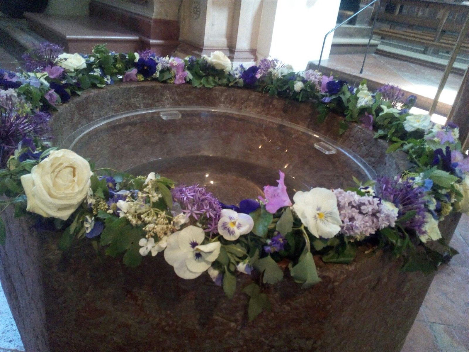 Blütenkranz aus Rosen, Veilchen und Flieder rund um einen steinernen Taufstein